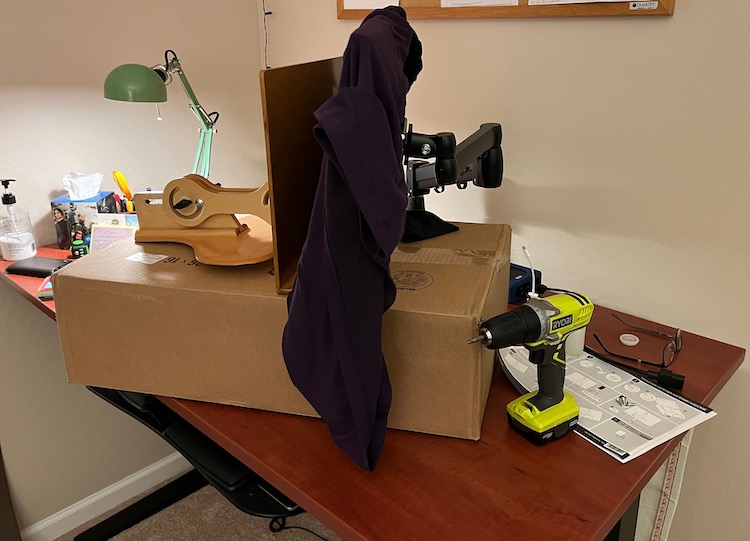 Photo of the top of desk. A cardboard box supports a wooden laptop stand that is holding a monitor vertical so the monitor arm can be attached. A drill is also visible.